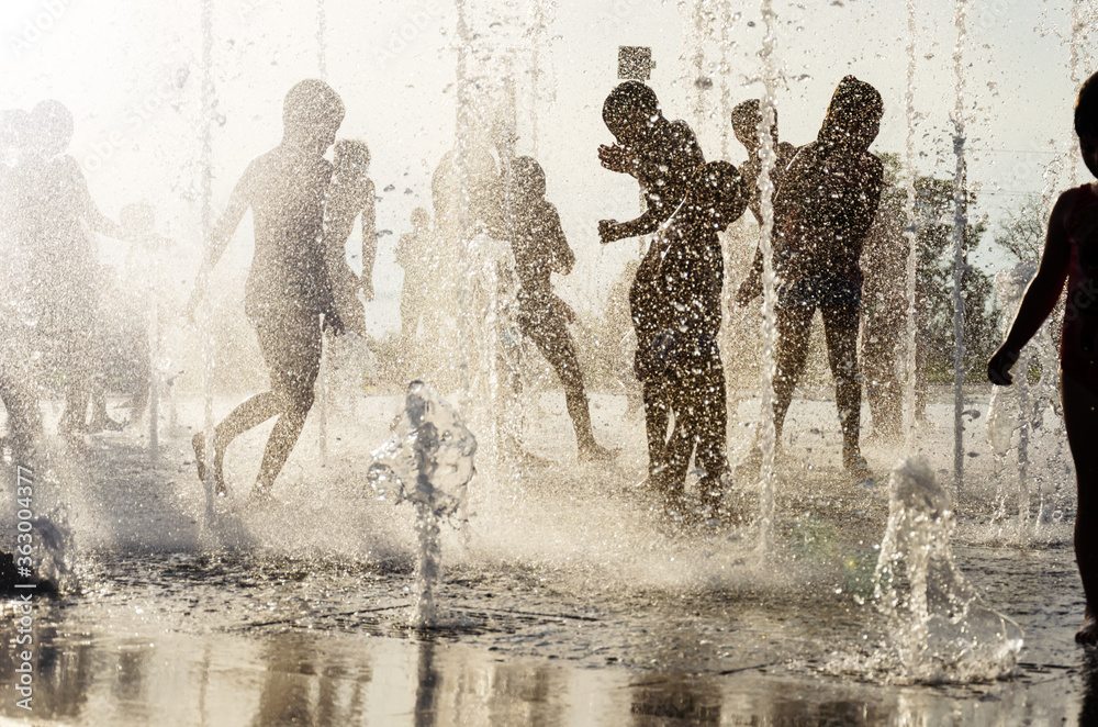Silhouettes of joyful children playing in water fountain on hot summer day. Happy childhood concept. Back light.