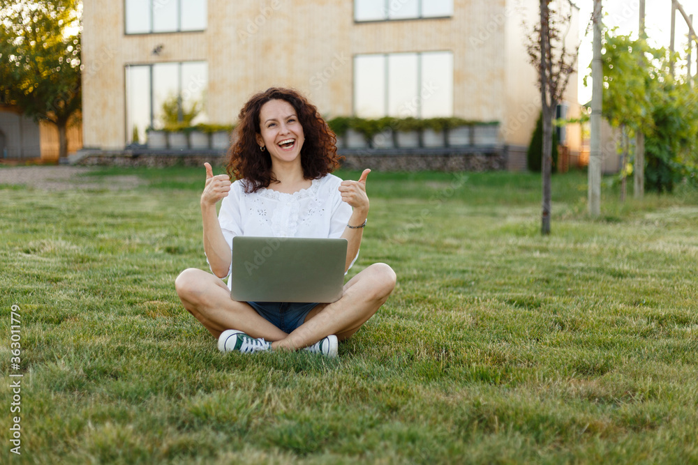 Frontal view of cheerful young woman sitting on green grass with legs crossed during summer day while using laptop smiling, holding thumbs up.