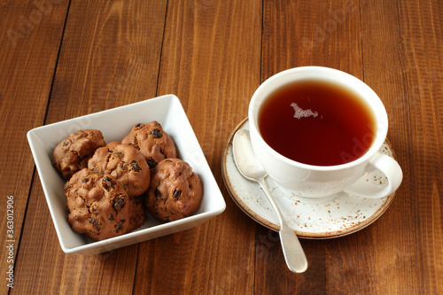 Chocolate cookies with walnuts and raisins in a square bowl and a cup of black tea on a wooden table. Closeup