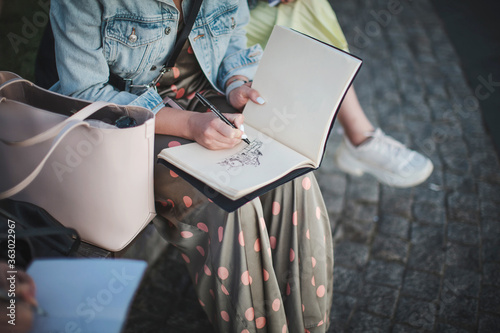 Young beautiful girl draws in a park in a group. Outline in a sketchbook.