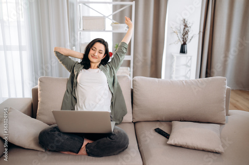 Young stylish girl is resting from using a laptop while sitting on a sofa in her cozy apartment