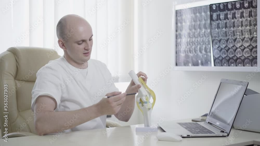 Portrait of positive professional doctor explaining joint structure on model and talking. Confident Caucasian physician or traumatologist sitting in hospital office. Job, occupation, lifestyle. Portrait of positive professional doctor explaining joint structure on model and talking. Confident Caucasian physician or traumatologist sitting in hospital office. Job, occupation, lifestyle.