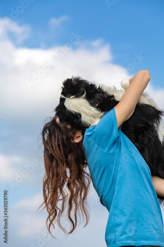 A girl with long hair in a blue T-shirt holds a black and white border collie dog in her arms. Portrait against a bright blue sky with clouds. Vertical orientation. 