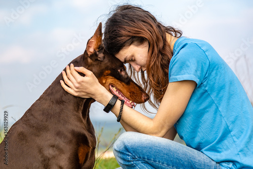 A girl in a blue T-shirt and a brown-and-tan doberman dobermann dog sit forehead to forehead against the sky. Horizontal orientation. 