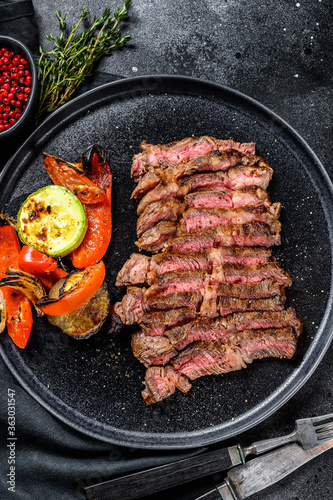 Sliced Grilled beef marbled rare steak. Chuck eye roll on a plate with a side dish of vegetables. Black background. Top view