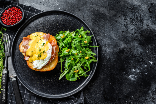 Brioche sandwich with bacon, egg Benedict and hollandaise sauce. Garnish with arugula salad. Black background. Top view. Copy space