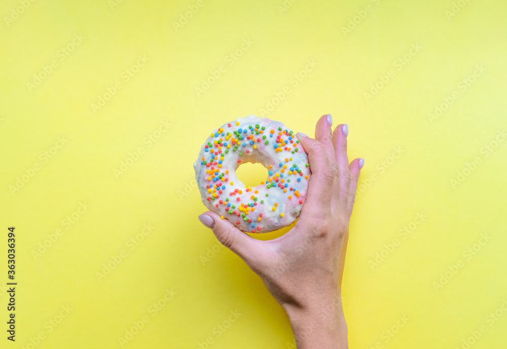 One donut with white icing. An overhead view of a doughnut with colored sprinkles. Female hand holding donut over yellow background. Top view, flat lay. Sweet, dessert, diet concept. One donut with white icing. An overhead view of a doughnut with colored sprinkles. Female hand holding donut over yellow background. Top view, flat lay. Sweet, dessert, diet concept.