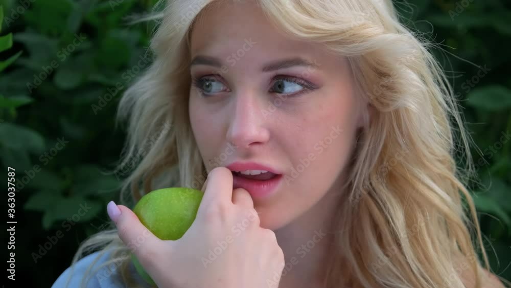 Portrait of a young beautiful woman with curly blond hair against a park and green trees. Beautiful face, blue eyes and luscious lips. The girl laughs playfully and holds a green apple in her hands. Portrait of a young beautiful woman with curly blond hair against a park and green trees. Beautiful face, blue eyes and luscious lips. The girl laughs playfully and holds a green apple in her hands.
