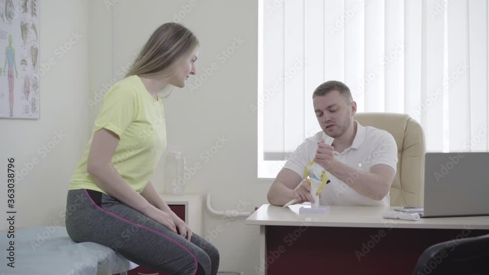Side view of brunette young woman sitting in doctor office. Professional Caucasian doctor showing knee joint model and talking to female patient. Traumatology, healing, treatment, medicine. Side view of brunette young woman sitting in doctor office. Professional Caucasian doctor showing knee joint model and talking to female patient. Traumatology, healing, treatment, medicine.
