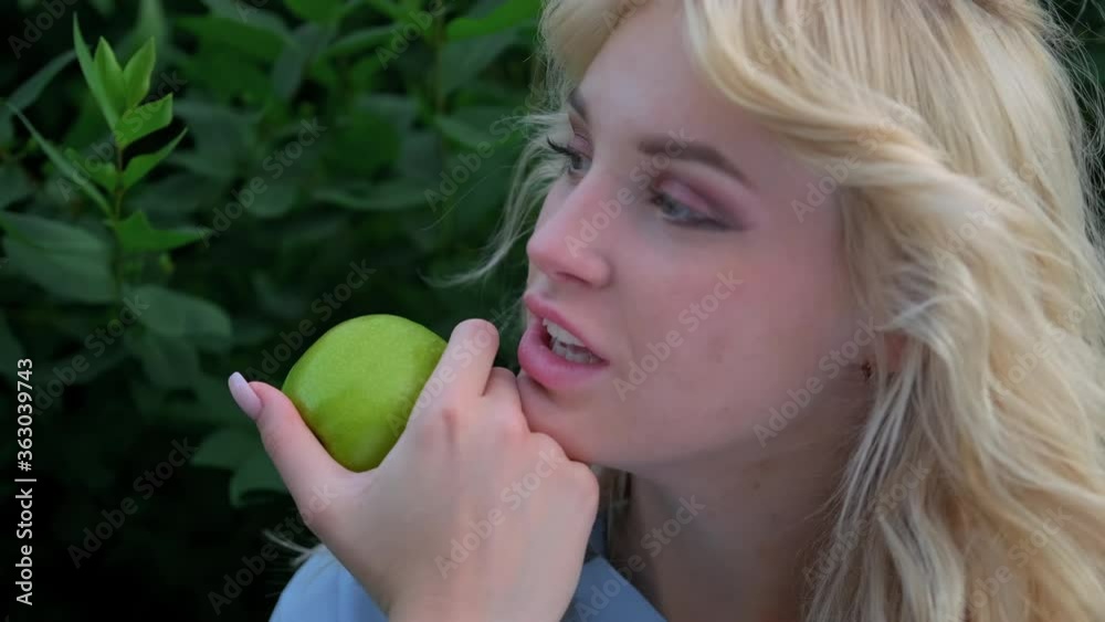 Portrait of a young beautiful woman with curly blond hair against a park and green trees. Beautiful face, blue eyes and luscious lips. The girl laughs playfully and holds a green apple in her hands. Portrait of a young beautiful woman with curly blond hair against a park and green trees. Beautiful face, blue eyes and luscious lips. The girl laughs playfully and holds a green apple in her hands.