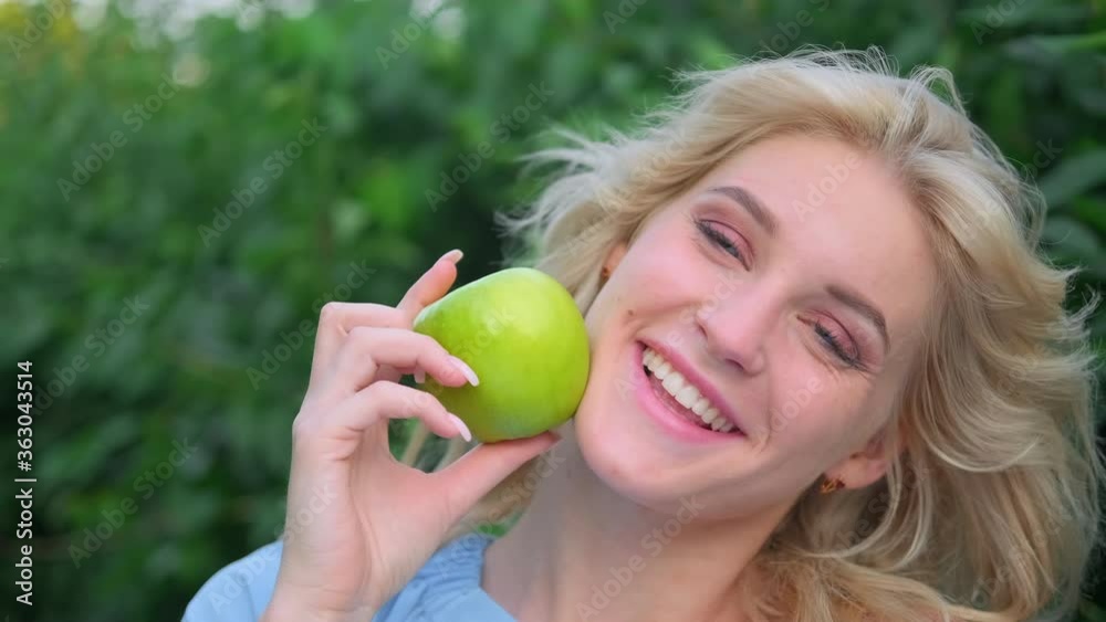 Portrait of a young beautiful woman with curly blond hair against a park and green trees. Beautiful face, blue eyes and luscious lips. The girl laughs playfully and holds a green apple in her hands. Portrait of a young beautiful woman with curly blond hair against a park and green trees. Beautiful face, blue eyes and luscious lips. The girl laughs playfully and holds a green apple in her hands.