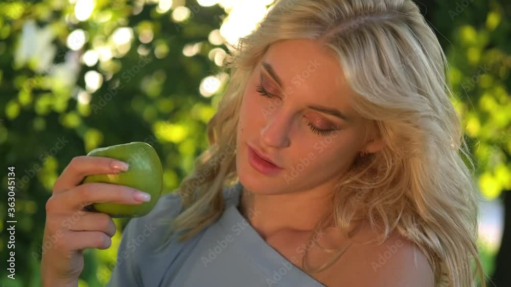 Portrait of a young beautiful woman with curly blond hair against a park and green trees. Beautiful face, blue eyes and luscious lips. The girl laughs playfully and holds a green apple in her hands. Portrait of a young beautiful woman with curly blond hair against a park and green trees. Beautiful face, blue eyes and luscious lips. The girl laughs playfully and holds a green apple in her hands.