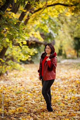 Autumn mood. Beautiful middle-aged woman weared in red jacket is smiling and walking in a park on background of beautifull autumn leaves. Fall coming. Autumn story. Yellow leaves in park. 
