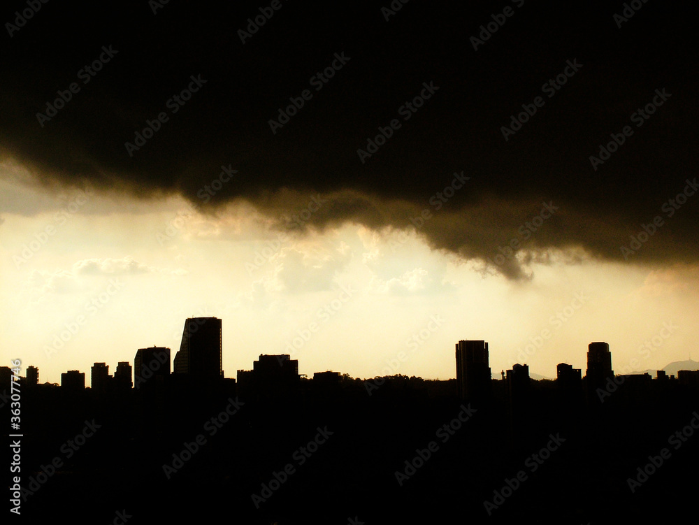 A silhueta de uma linha de prédios em São Paulo e uma escura nuvem de tempestade encobrindo o céu da cidade no meio da tarde.