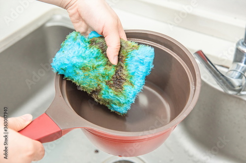 A hand in front of the sink with a blue cleaning cloth, cleaning a greasy soup pot. 