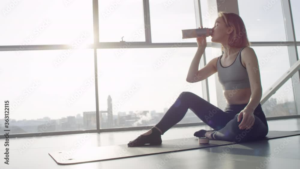 Attractive young woman sitting relaxed on yoga mat in modern gym drinking fresh water during break time