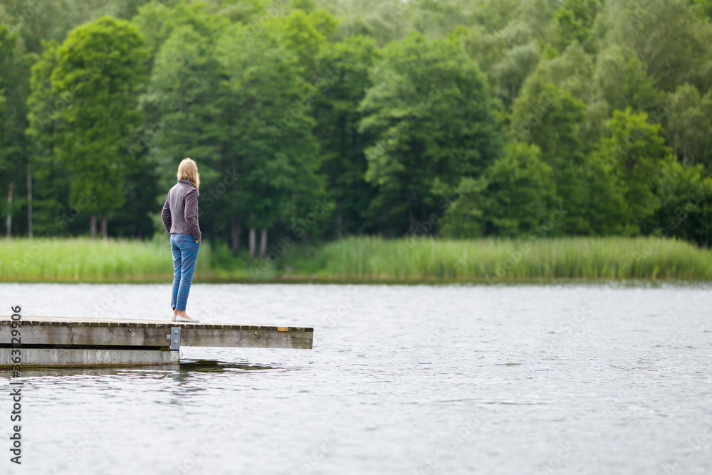 One young adult woman standing alone on edge of footbridge and staring at lake and green trees in summer day. Thinking about life. Spending time alone in nature. Peaceful atmosphere. Back side view.