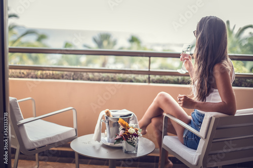 Young woman enjoying vacation at resort. Relaxing at balcony with champagne glass, looking at view. (Soft focus)
