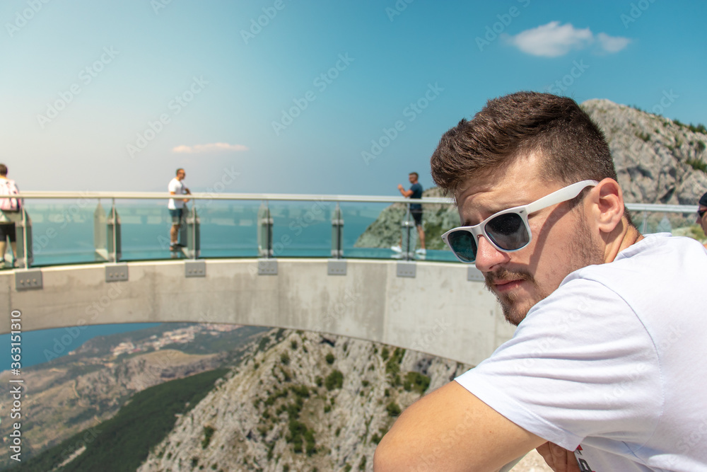 Man standing on the entrance of the skywalk going accross the biokovo mountain, looking into the distance. People seen walking accross the glass structure in the background