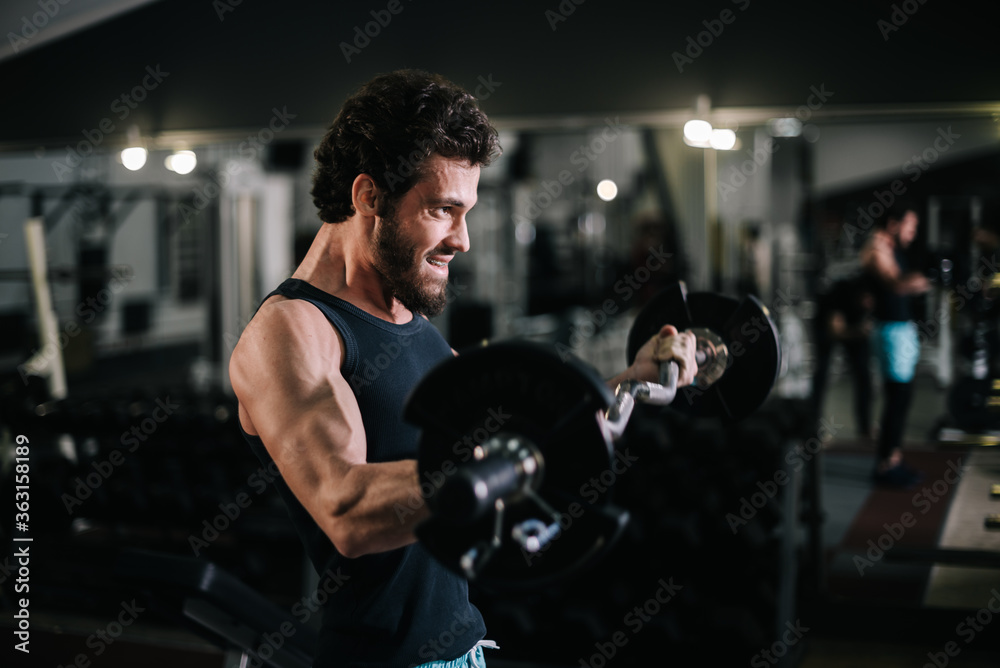 Side view of sportive bearded young man with muscular wiry body wearing sportswear working out with barbell during sport workout training in modern dark gym. Concept of healthy lifestyle. Side view of sportive bearded young man with muscular wiry body wearing sportswear working out with barbell during sport workout training in modern dark gym. Concept of healthy lifestyle.