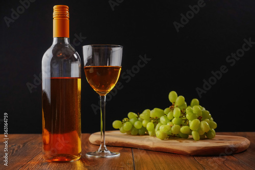 Stil life with white jucy grapes, bottle of white wine without any label, and one wine glass. Dark black background, Wooden table.