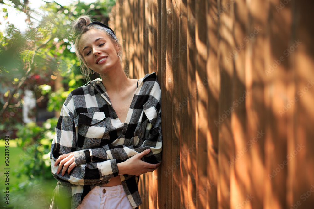 Portrait of a pretty girl in a plaid shirt and bandana standing against a wooden fence in the backyard on a Sunny day.