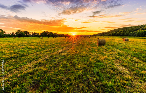 Scenic view at beautiful sunset in a green shiny field with hay stacks, bright cloudy sky , trees and golden sun rays with glow, summer valley landscape