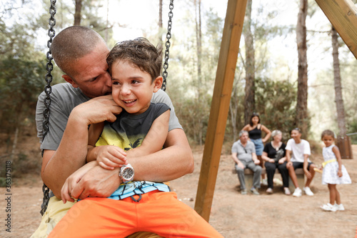 papa gioca con il figlio seduti su un altalena e solo sfondo il resto della famiglia in un parco giochi in un bosco photo