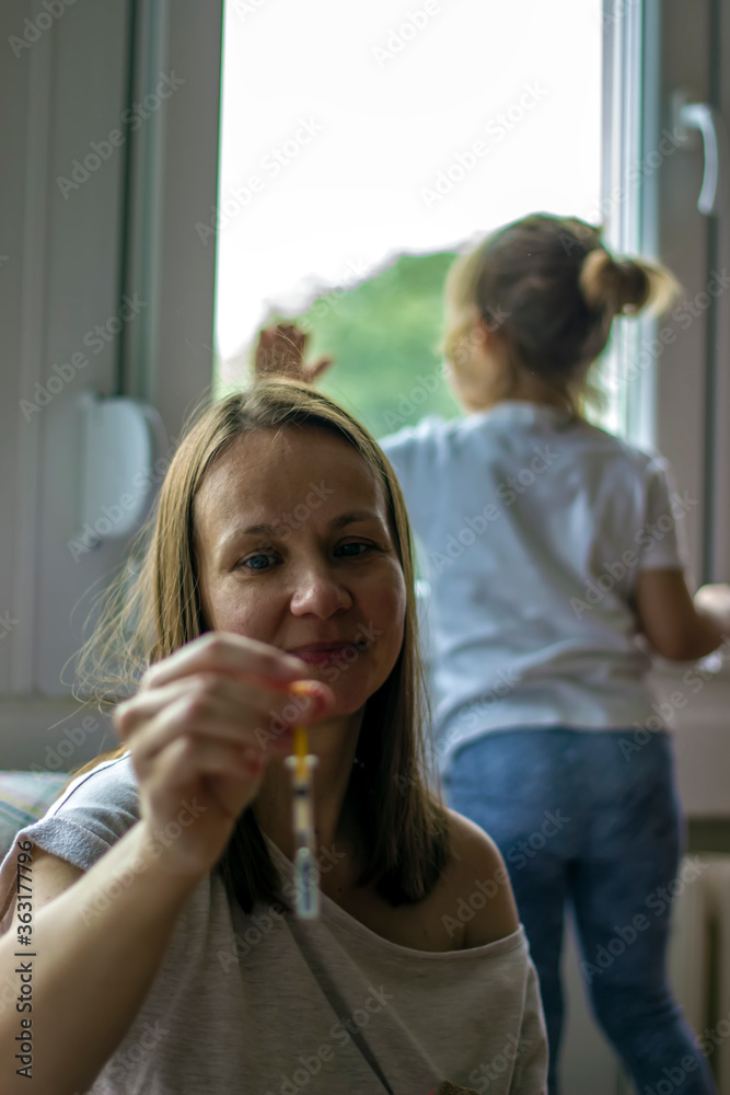Mother is showing syringe while daughter is looking through window.Shot of young mother holding her medication.Cute girl turned back to her mother,looking out the window,while mother takes medication Mother is showing syringe while daughter is looking through window.Shot of young mother holding her medication.Cute girl turned back to her mother,looking out the window,while mother takes medication