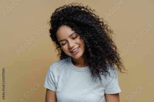 Photo of pleased satisfied woman smiles with pleasure, closes eyes and shows snow white teeth, has curly hairstyle, wears casual t shirt, poses against beige background. Happiness, joy concept