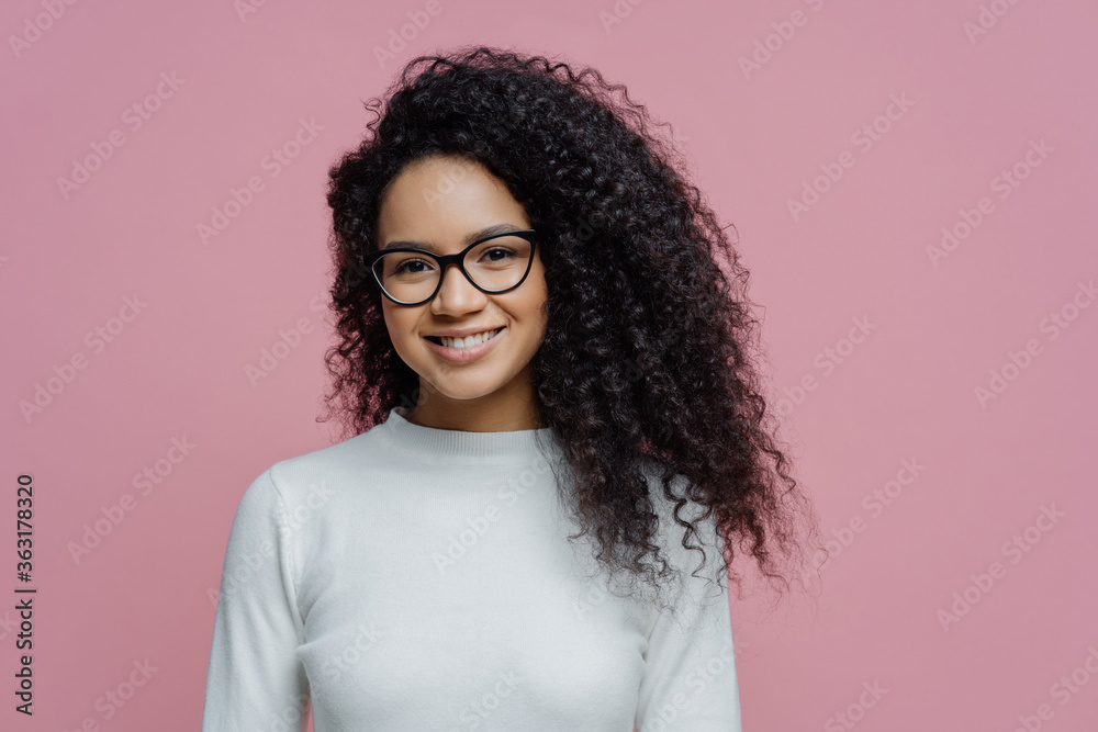 Headshot of lovely curly haired woman with healthy skin and toothy smile, dressed in white jumper, wears transparent glasses, isolated over rosy background. People, face expressions and emotions Headshot of lovely curly haired woman with healthy skin and toothy smile, dressed in white jumper, wears transparent glasses, isolated over rosy background. People, face expressions and emotions