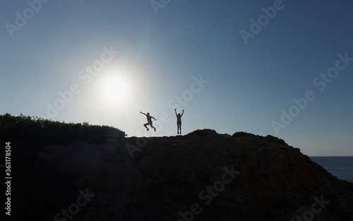 Silhouette of two people jumping happy, on top of a mountain, at sunset.