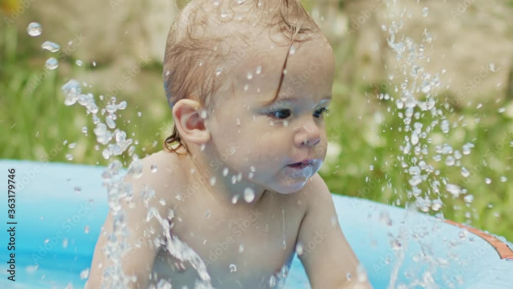 Cute little child bathing in blue street pool in courtyard. Portrait of joyful toddler, baby. Kid laughs, splashes water, smiles. Concept of healthy lifestyle, family, leisure in summer. Slow motion.