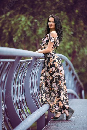 A beautiful girl stands near the fence on the bridge. Beautiful girl on a blurred background of nature. Beautiful girl in a long dress. Everyday life of a beautiful young lady.