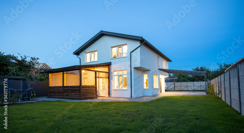 Modern minimalist white color house exterior in the evening with garden, lawn and patio area in summer time
