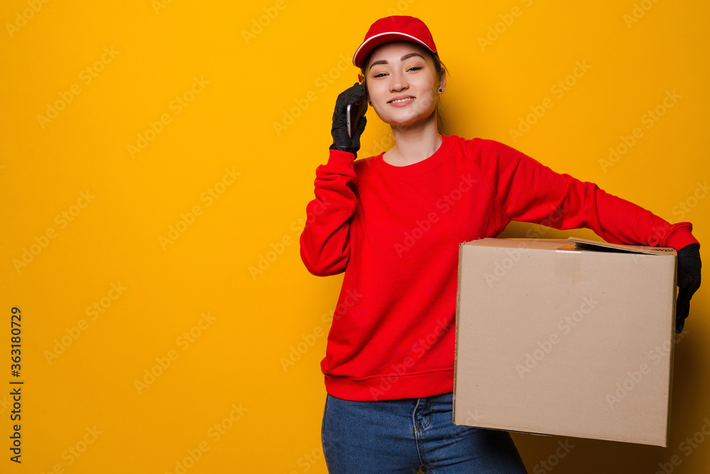Young asian delivery woman holding box talking on the phone isolated on yellow background Young asian delivery woman holding box talking on the phone isolated on yellow background
