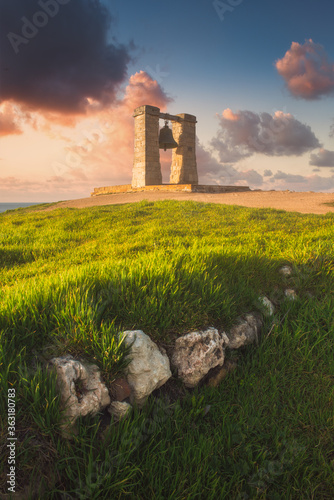 Picturesque landscape with an ancient foggy bell in the center of the frame. Spring evening by the sea, magical sunset, ancient masonry, fresh grass and foggy bell. Copy space on the colorful sky. 