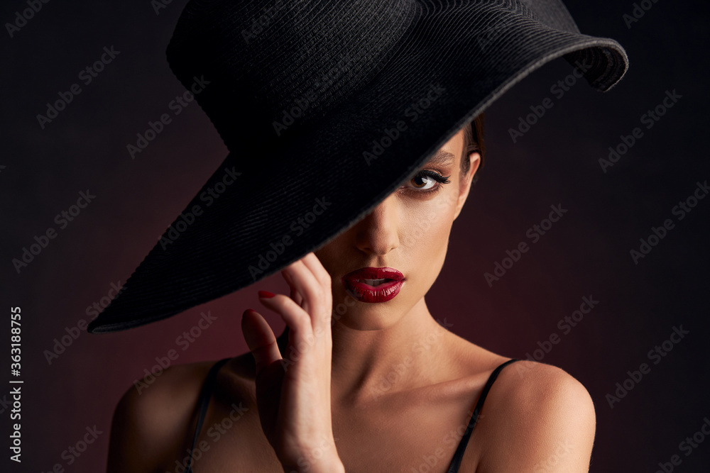 portrait of beautiful tanned girl with professional makeup, red lips, on a burgundy background in a black dress with straps and black hat that covers her eye she looks at the camera with serious face portrait of beautiful tanned girl with professional makeup, red lips, on a burgundy background in a black dress with straps and black hat that covers her eye she looks at the camera with serious face