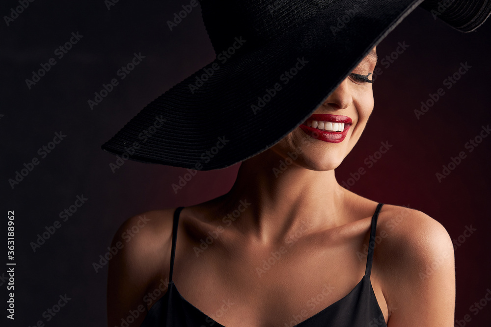 portrait of a beautiful tanned girl with professional makeup on a burgundy background in a black dress with straps, black hat that covers her eye, she smiles with white teeth portrait of a beautiful tanned girl with professional makeup on a burgundy background in a black dress with straps, black hat that covers her eye, she smiles with white teeth