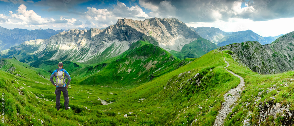 Hiker Man on Hiking Trail with Backpack standing relaxed and enjoying panoramic view to mountains. Alps, Hohe Gaenge, Allgau, Bad Hindelang, Hinterstein, Bavaria, Germany. - obrazy, fototapety, plakaty 