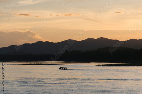 The boat sailed on the river with the setting sun setting, with a mountain view in the background, the reflections from the river in orange.