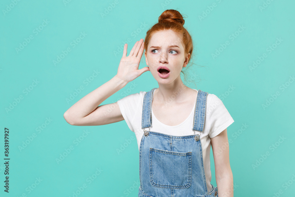 Shocked young readhead girl in casual denim clothes posing isolated on blue turquoise wall background studio portrait. People lifestyle concept. Mock up copy space. Try to hear you listening intently.
