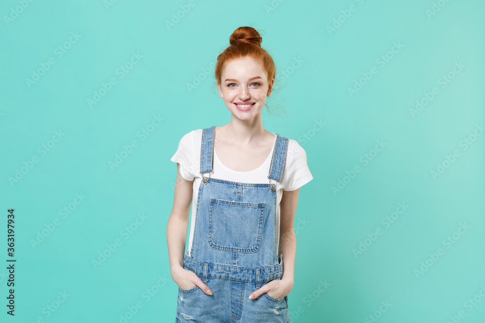 Smiling young readhead girl in casual denim clothes isolated on blue turquoise wall background studio portrait. People sincere emotions lifestyle concept. Mock up copy space. Holding hands in pockets. Smiling young readhead girl in casual denim clothes isolated on blue turquoise wall background studio portrait. People sincere emotions lifestyle concept. Mock up copy space. Holding hands in pockets.