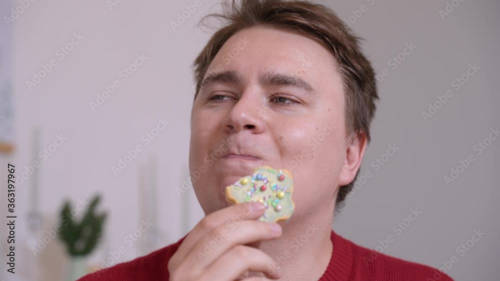 Young plump man bites off a piece of cookie in the shape of a Christmas tree, intricately decorated, eats with great pleasure and makes an approving gesture with his finger. Home-made cooking concept Young plump man bites off a piece of cookie in the shape of a Christmas tree, intricately decorated, eats with great pleasure and makes an approving gesture with his finger. Home-made cooking concept