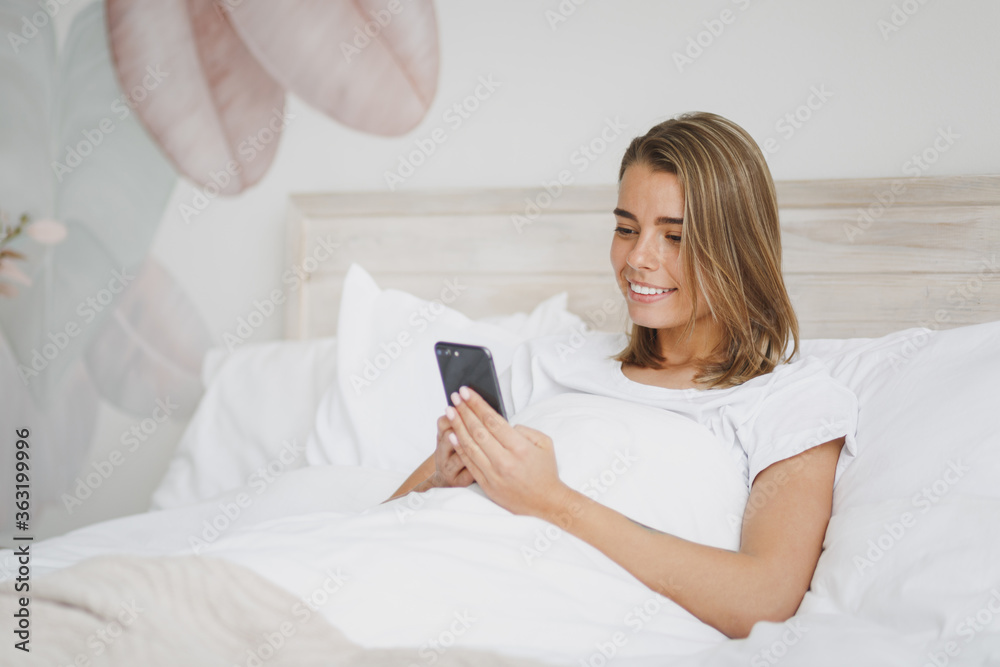 Beautiful young girl lying in bed with white sheet pillow blanket spending time in bedroom at home. Rest relax good mood lifestyle concept. Mock up copy space. Using mobile phone, typing sms message. Beautiful young girl lying in bed with white sheet pillow blanket spending time in bedroom at home. Rest relax good mood lifestyle concept. Mock up copy space. Using mobile phone, typing sms message.