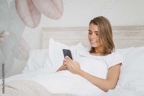 Beautiful young girl lying in bed with white sheet pillow blanket spending time in bedroom at home. Rest relax good mood lifestyle concept. Mock up copy space. Using mobile phone, typing sms message.