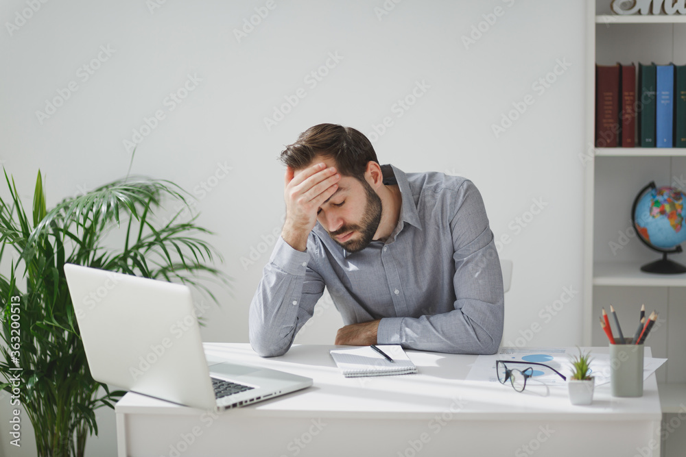 Exhausted tired business man in gray shirt sitting at desk work on laptop pc computer in light office on white wall background. Achievement business career concept. Put hand on head having headache. Exhausted tired business man in gray shirt sitting at desk work on laptop pc computer in light office on white wall background. Achievement business career concept. Put hand on head having headache.