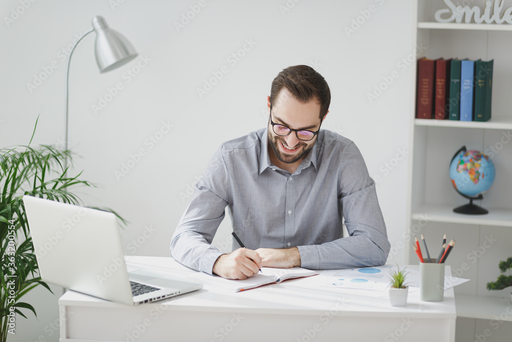 Smiling young bearded business man in gray shirt glasses sit at desk work on laptop pc computer in light office on white wall background. Achievement business career concept Writing notes in notebook. Smiling young bearded business man in gray shirt glasses sit at desk work on laptop pc computer in light office on white wall background. Achievement business career concept Writing notes in notebook.