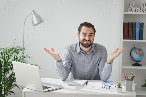 Perplexed puzzled young bearded business man in gray shirt sitting at desk work on laptop pc computer in light office on white wall background. Achievement business career concept. Spreading hands.