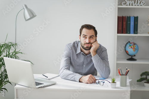 Boring young bearded business man in gray shirt sitting at desk working on laptop pc computer in light office on white wall background. Achievement business career concept. Put hand prop up on cheek. photo
